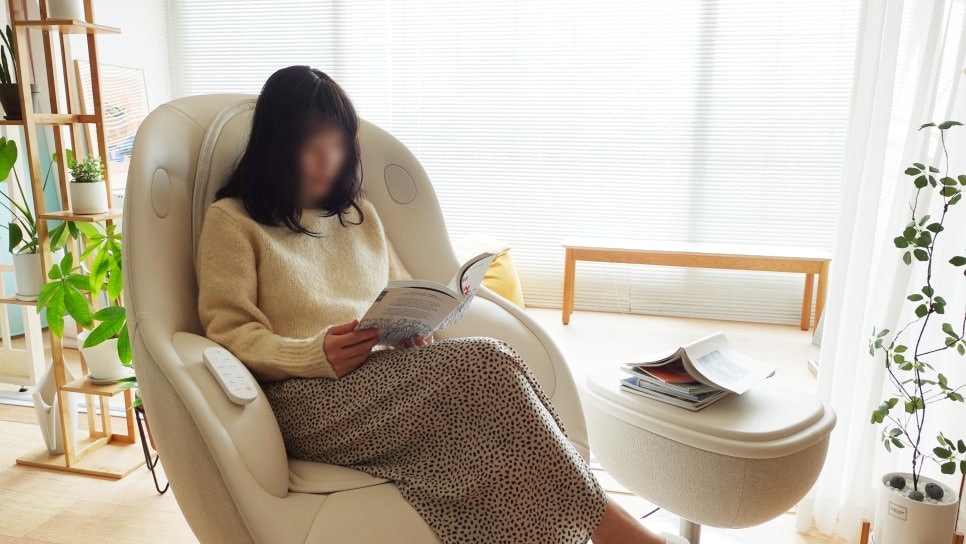 한_사람이_안마의자에_앉아_책을_읽고_있음._옆에는_발받침_위에_잡지가_놓여_있으며,_배경에는_블라인드로_빛이_은은하게_들어오는_창과_실내_화초,_우드_벤치가_배치됨._아늑하고_편안한_분위기의_공간임.