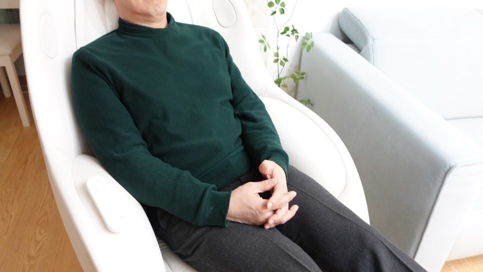 한_사람이_안마의자에_편안하게_기대어_앉아_있음._팔걸이에는_내장_리모컨이_배치되어_있으며,_배경에는_블루톤_소파와_실내_화초가_놓여_있음._아늑하고_편안한_분위기의_공간임.