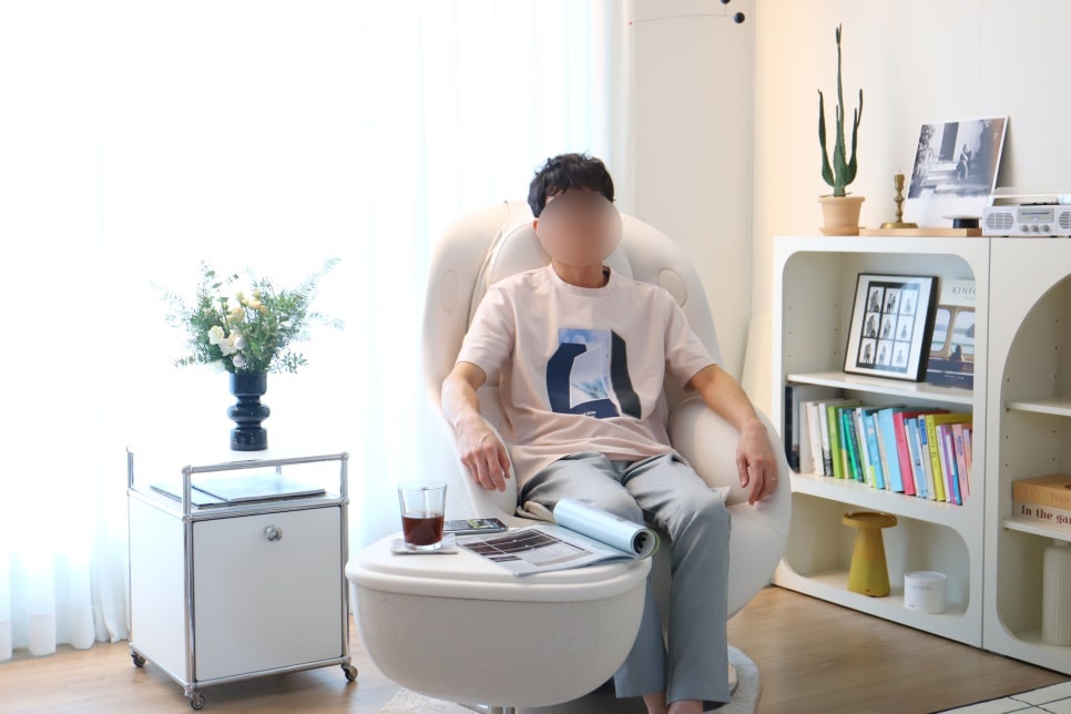한_사람이_흰색_안마의자에_편하게_기대어_있고,_발받침_테이블에는_잡지와_커피잔이_놓여_있음._옆엔_검은색_꽃_화병이_있는_금속_선반과_선인장,_책이_정리된_흰_선반장이_있음._뒤로는_채광이_들어오는_커튼이_보임.