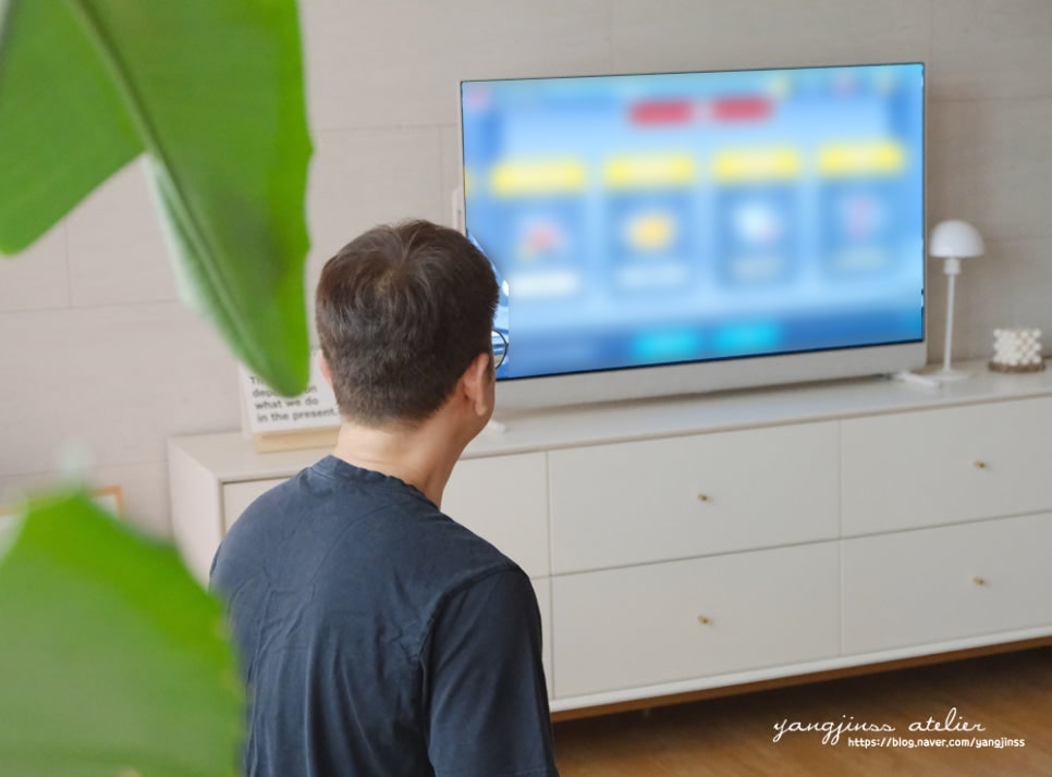 거실에서_남성이_tv_화면을_바라보고_있음._tv는_수납장_위에_놓여_있으며,_옆에는_스탠드_조명과_장식_소품이_배치됨.