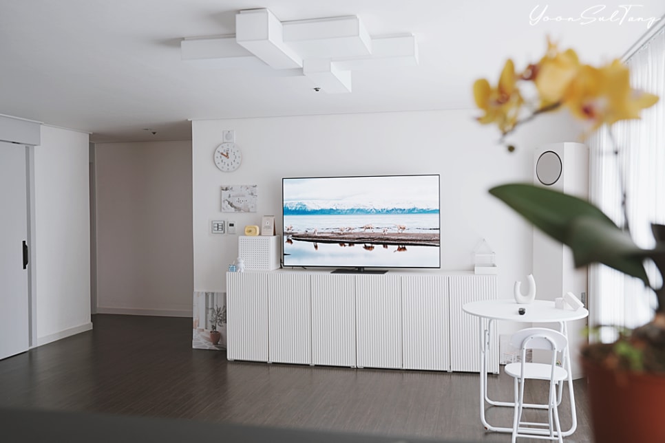 거실에_흰색_수납장_위에_tv가_놓여_있음._tv_화면에는_호숫가_풍경이_표시됨._배경에는_벽걸이_시계,_액자,_원형_타워형_에어컨,_흰색_테이블과_의자가_있음._앞쪽에_노란_난초_화분이_있음.