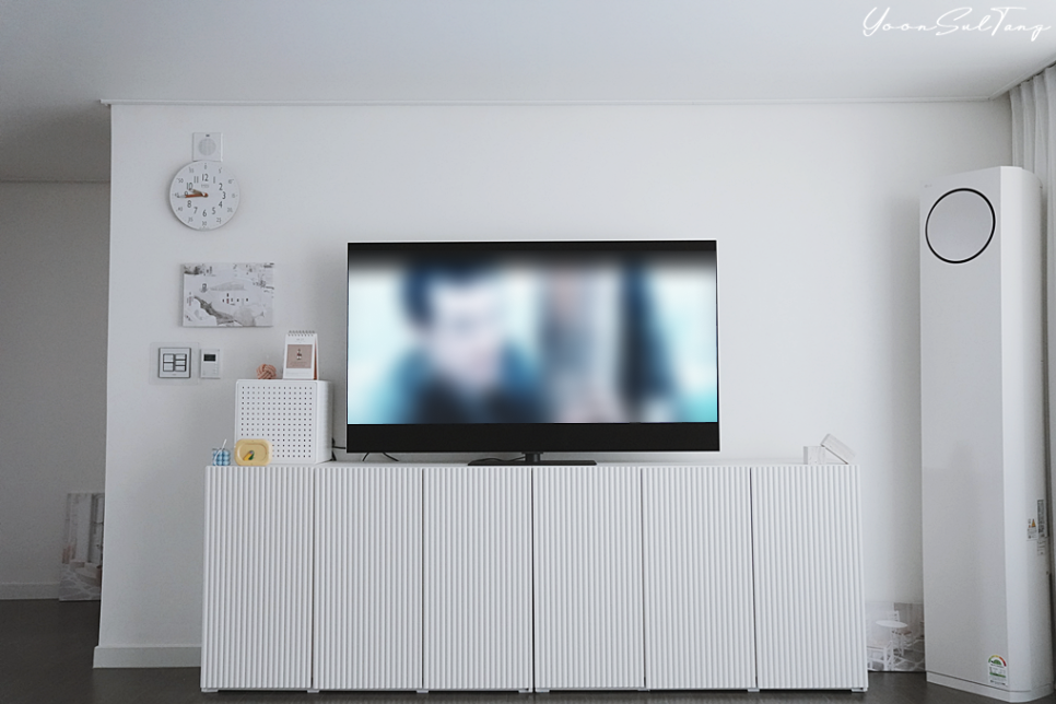 거실에_흰색_수납장_위에_tv가_놓여_있음._tv_화면에는_흐릿한_인물_영상이_표시됨._배경에는_벽걸이_시계,_액자,_원형_타워형_에어컨,_작은_장식품이_있음.