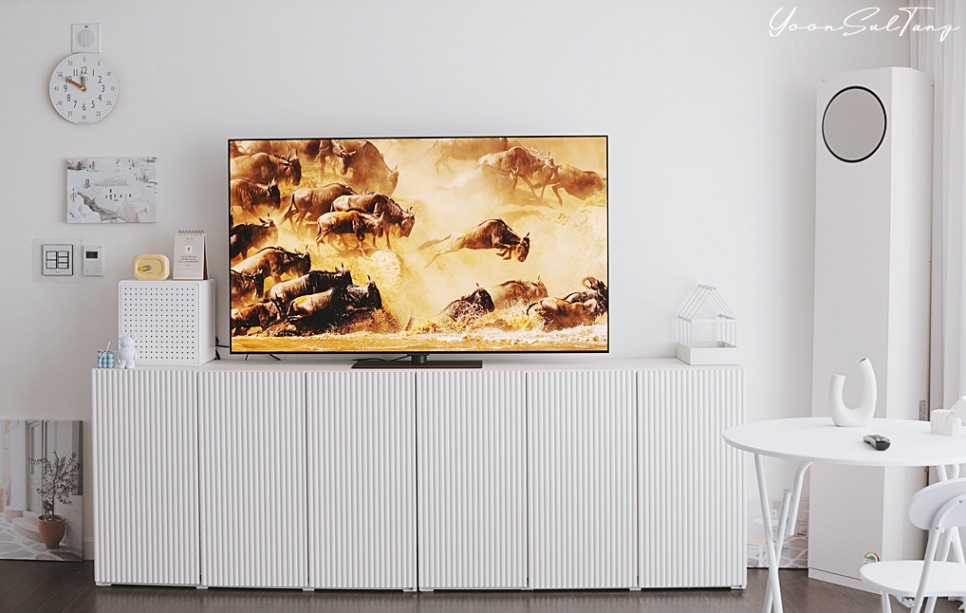 거실에_흰색_수납장_위에_tv가_놓여_있음._tv_화면에는_황토색_먼지를_일으키며_달리는_누_떼가_표시됨._배경에는_벽걸이_시계,_액자,_원형_타워형_에어컨,_흰색_테이블과_의자가_있음.
