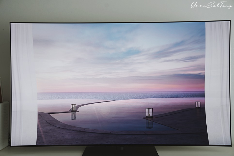거실에_놓인_대형_tv_화면에_양쪽에_커튼이_있고,_바다가_보이는_풍경이_표시됨.