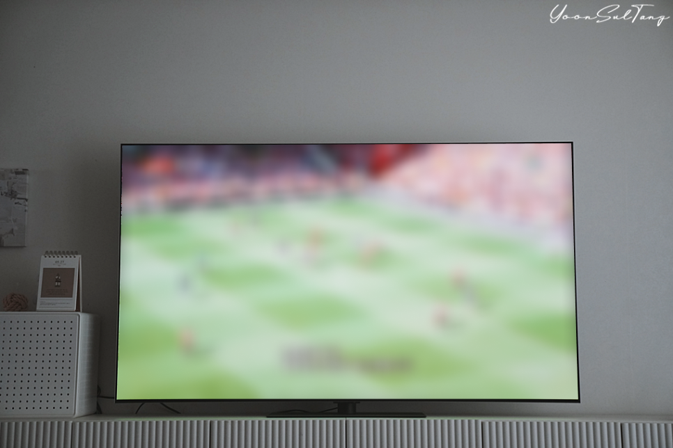 거실에_놓인_대형_tv_화면에_축구_경기가_흐림_효과로_표시됨._배경에는_장식장과_소품들이_있음.