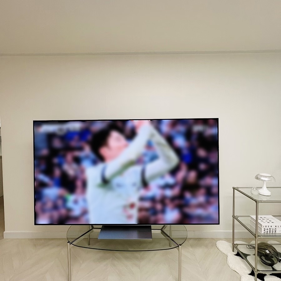거실_테이블_위_tv에서_스포츠_경기_영상이_재생_중임._옆에는_유리_선반과_장식품이_있음.