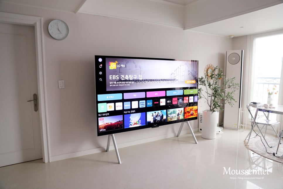거실에_설치된_lg_signature_oled_m_tv가_다양한_ott_애플리케이션과_콘텐츠를_표시하는_홈_화면을_보여주고_있다._tv는_얇은_베젤과_심플한_스탠드_디자인을_갖추고_있으며,_오른쪽에는_에어컨과_화분이_배치되어_있다.