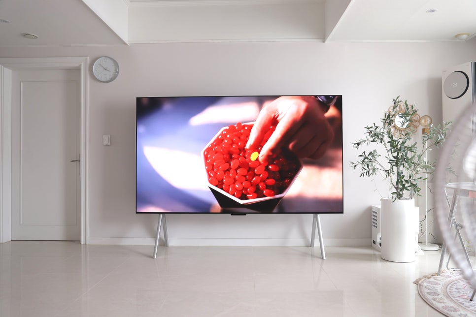lg_signature_oled_m_tv가_손으로_빨간_젤리캔디를_집어드는_영상을_재생하고_있다._화면의_밝고_선명한_색상이_강조되며,_tv가_거실_벽면에_세워져_있다._오른쪽에는_에어컨과_화분,_작은_테이블이_놓여_있다.