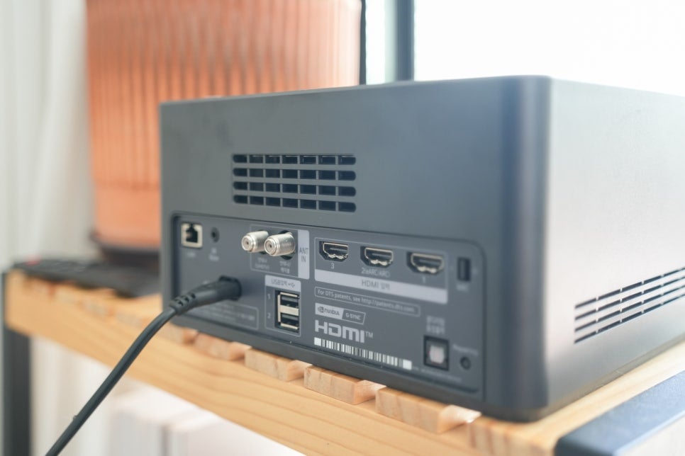 lg_signature_oled_m의_제로커넥터_박스_후면._나무_선반_위에_놓여_있으며,_hdmi_포트,_usb_포트,_안테나_입력_등의_다양한_단자가_보인다._기기는_깔끔한_블랙_디자인으로_제작되었으며,_여러_개의_케이블이_연결되어_있다.