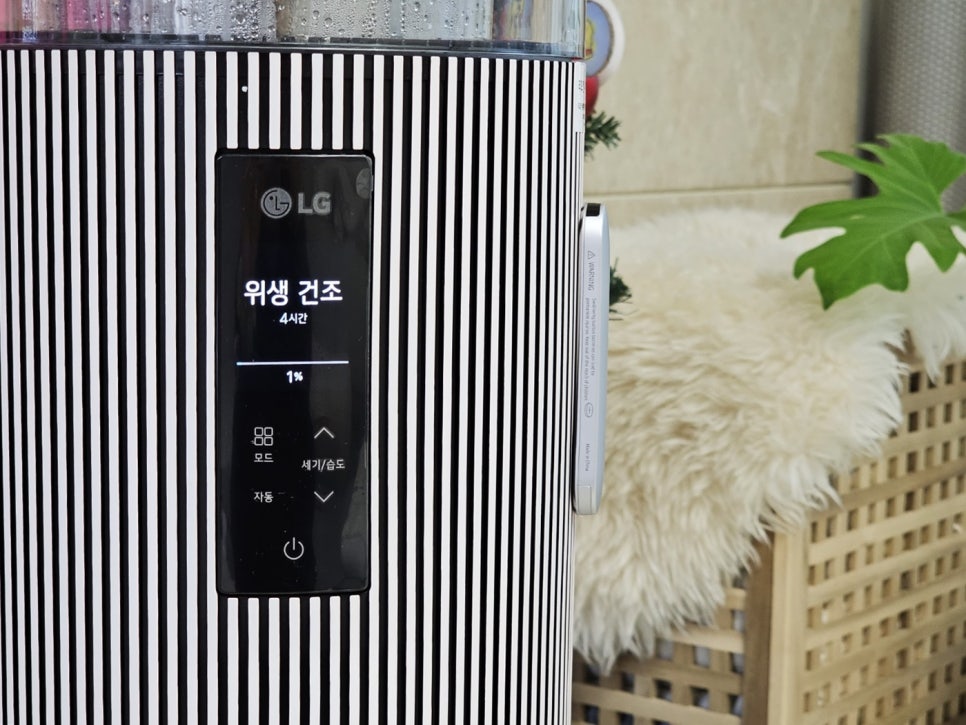 가까이에서_본_공기청정기의_디스플레이,_위생_건조_모드가_작동_중인_상태._세련된_스트라이프_패턴의_본체_디자인이_돋보이며,_배경에는_라탄_가구와_화분이_조화를_이루고_있음.