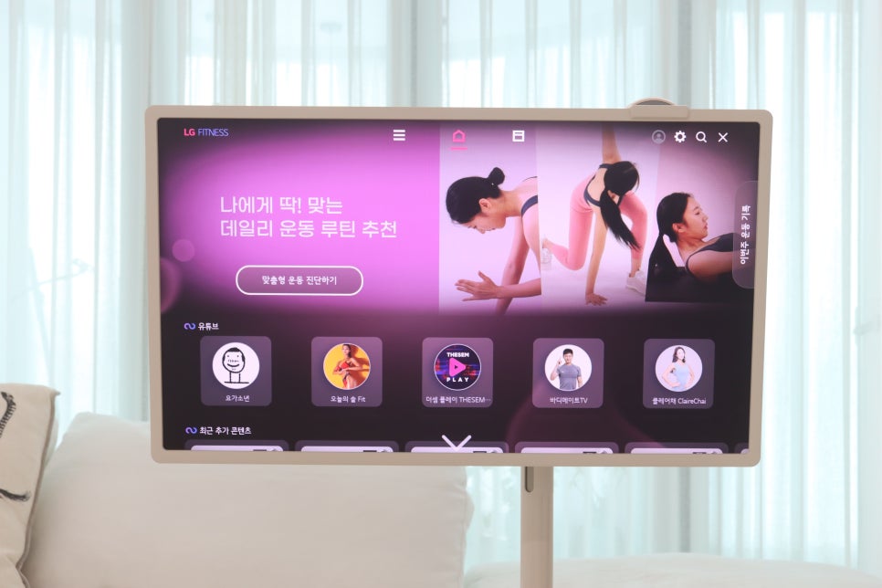 스탠바이미_화면에_lg_fitness_홈_화면_표시됨._개인_맞춤_운동_루틴_추천과_요가,_홈트레이닝_등_다양한_운동_콘텐츠_아이콘_있음._배경에는_소파와_밝은_커튼_있음.