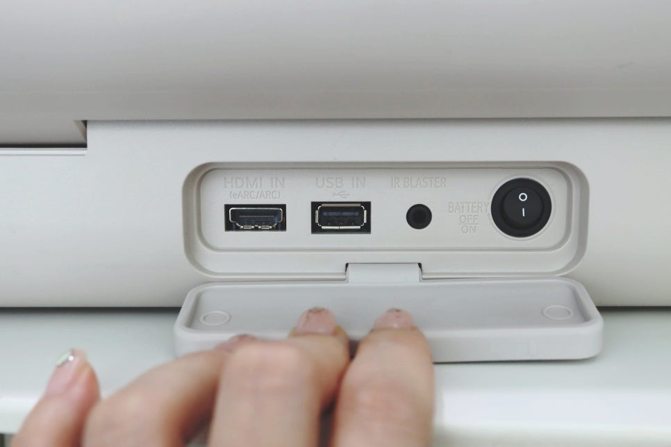 스탠바이미_go_후면_포트_덮개를_손으로_열고_있음._내부에는_hdmi_입력,_usb_입력,_ir_블라스터,_배터리_스위치,_전원_버튼_있음.