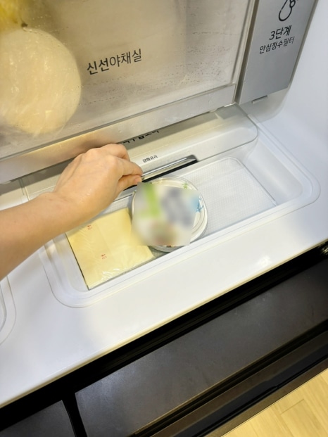 손이 냉장고 안의 3단계 안심살균필터가 있는 공간에서 작은 접시를 꺼내는 모습. 주변에는 식재료와 포장된 제품이 정리되어 있으며, 냉장고 내부가 깔끔하게 정리된 모습.