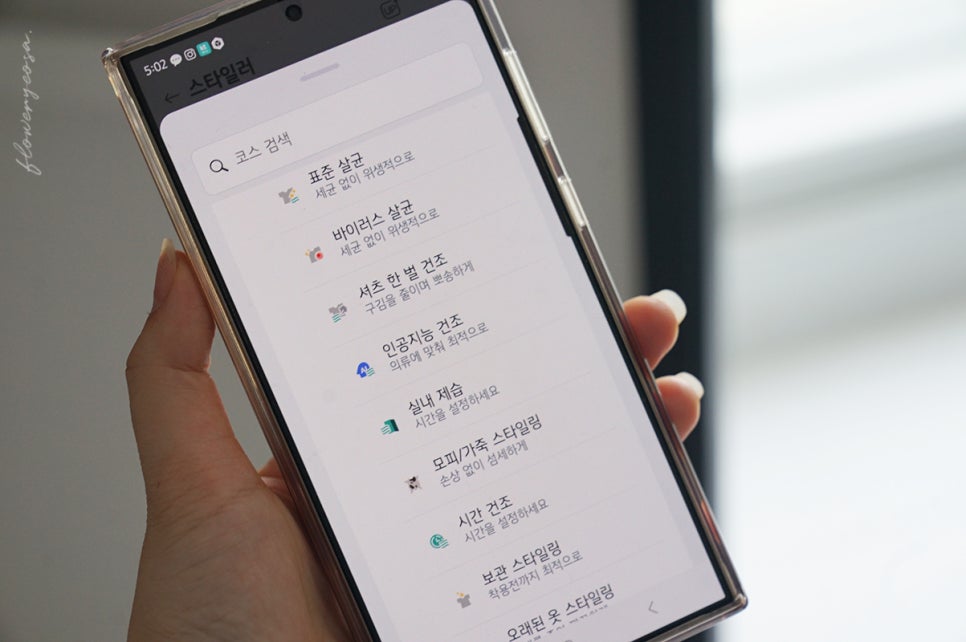 손에는_스타일러_전용_앱을_실행한_화면의_스마트폰을_들고_있음._표준_살균,_바이러스_살균,셔츠_한_벌_건조_등_다양한_관리_모드가_표시되어_있음.