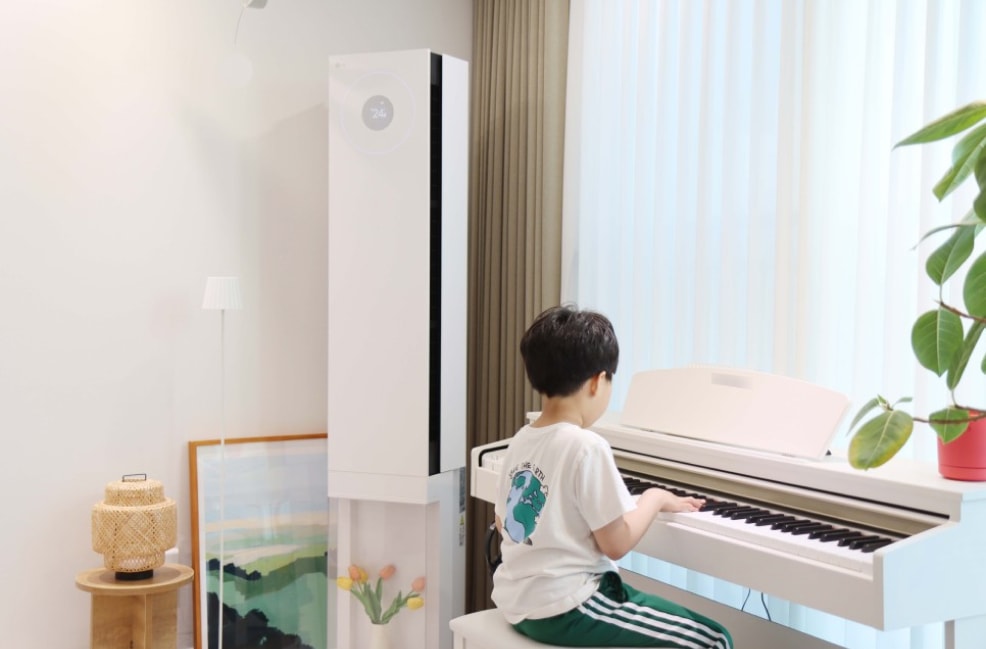 화이트_톤_거실에서_한_아이가_흰색_전자_피아노를_연주하고_있음._옆에는_스탠드_에어컨과_튤립이_담긴_장식_공간이_있으며,_원목_테이블과_라탄_조명,_그림_액자가_배치됨._배경에는_커튼과_초록_화분이_있음.
