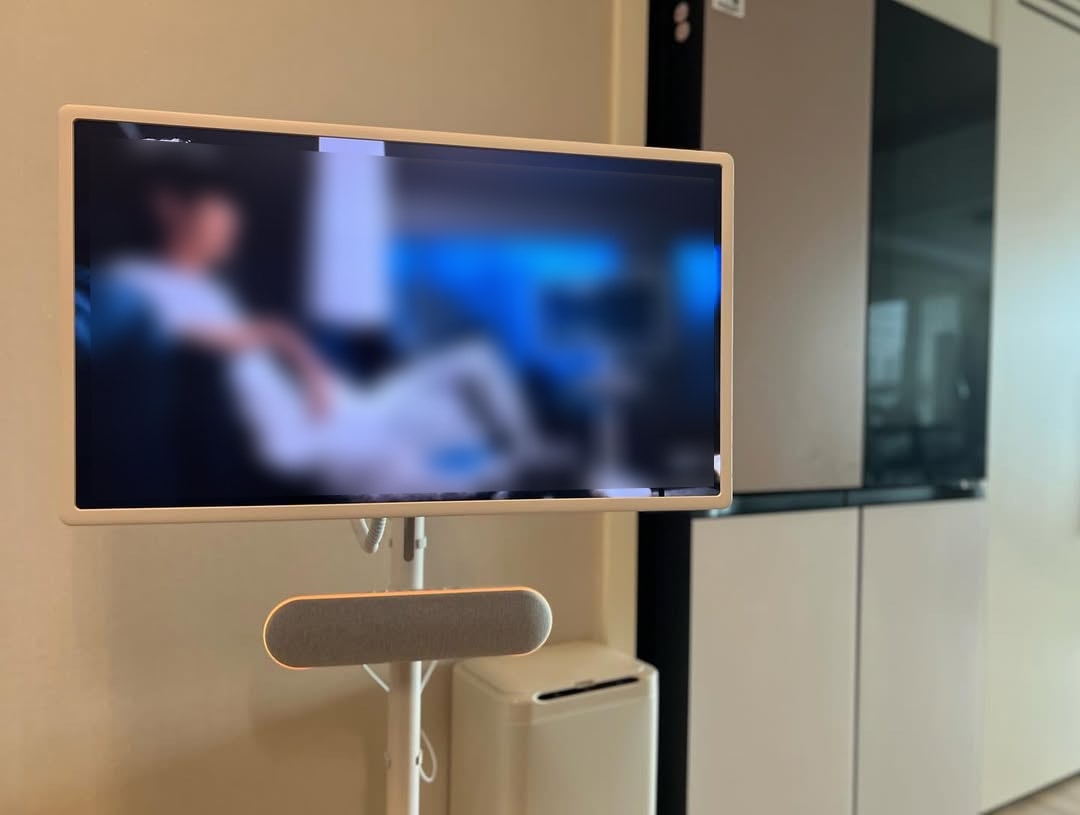 LG 스탠바이미 TV가 거실에 설치되어 있으며, 화면에는 영상이 재생되고 있다. TV는 스탠드형 디자인으로 스피커가 하단에 부착되어 있으며, 배경에는 LG 오브제컬렉션 냉장고와 가전제품이 놓여 있는 주방 공간이 보인다.