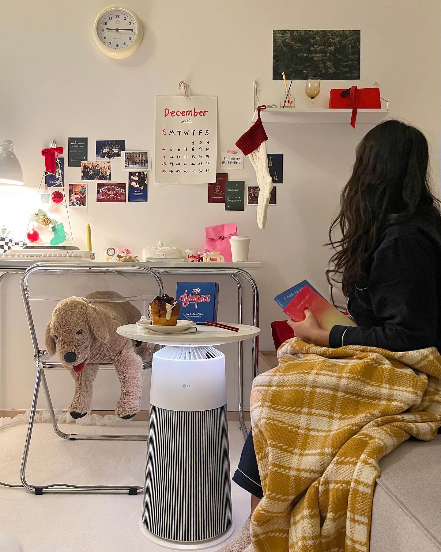 벽에 달력과 장식이 꾸며진 방에서 노란 체크 담요를 두른 사람이 책을 들고 있으며, LG 에어로퍼니처 테이블 위에는 디저트와 책이 놓여 있는 모습
