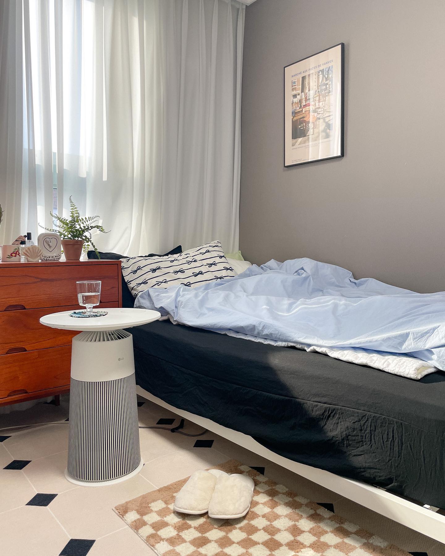 편안하고 밝은 침실, LG 공기청정기를 사이드 테이블로 사용하여 물컵과 소품이 놓여 있으며, 침대 옆에는 부드러운 슬리퍼와 나무 서랍장이 배치된 아늑한 공간