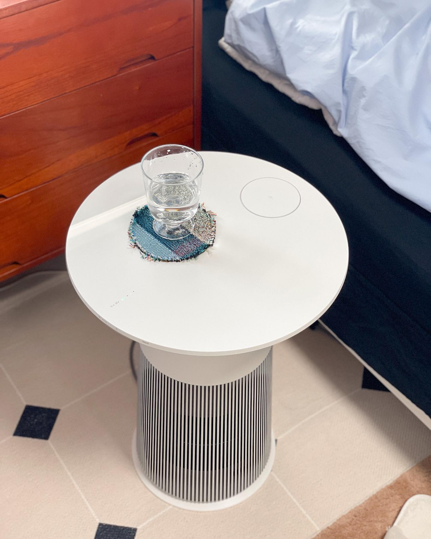 LG 공기청정기를 사이드 테이블로 활용하여 물컵이 놓여 있는 깔끔한 침실 공간, 나무 서랍장과 체크 패턴의 러그가 조화로운 아늑한 분위기