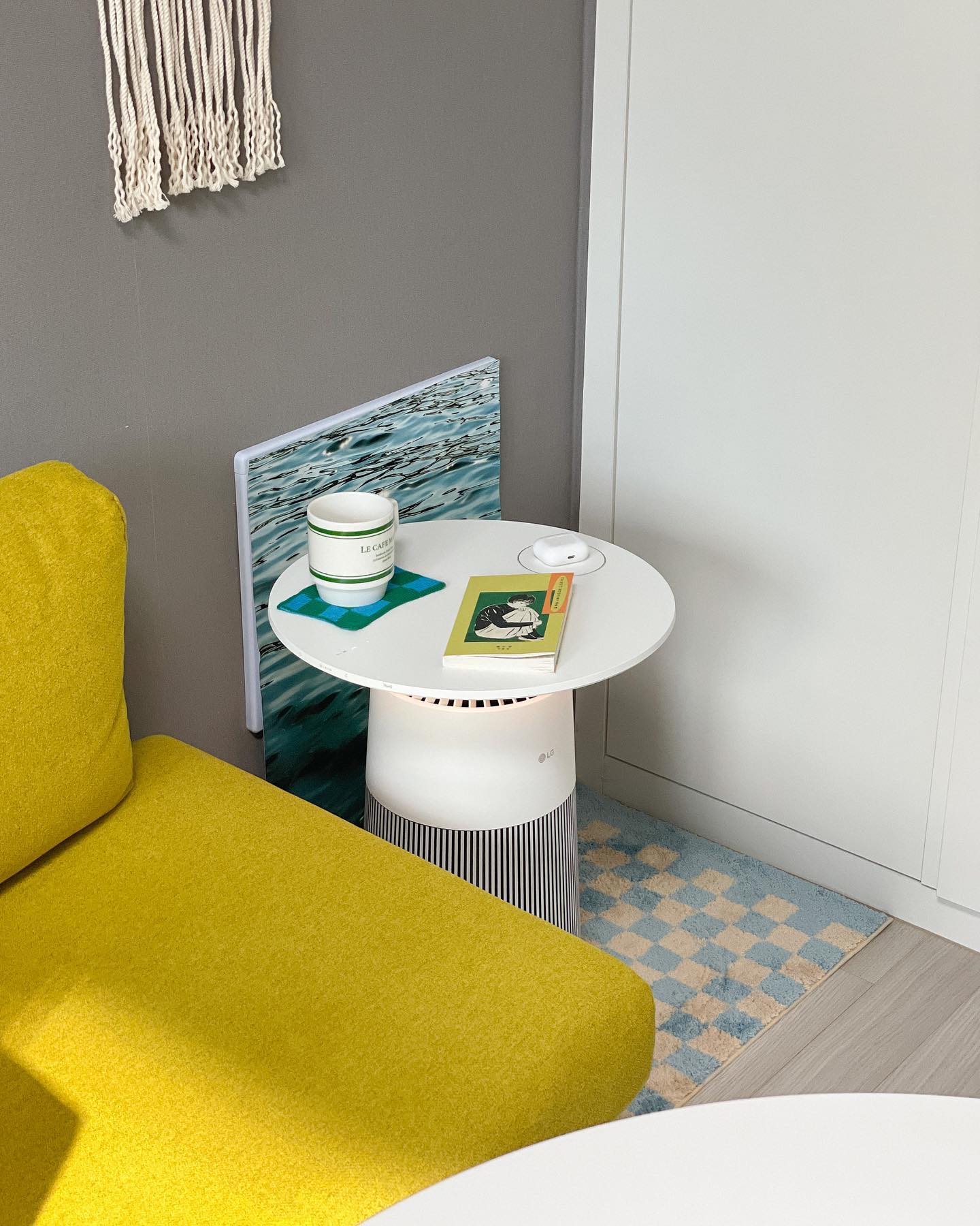 밝고 색감이 돋보이는 거실, LG 공기청정기를 사이드 테이블로 사용하여 머그컵, 책, 무선 이어폰이 놓여 있으며, 노란색 소파와 체크 패턴 러그가 아늑한 분위기를 더하는 공간
