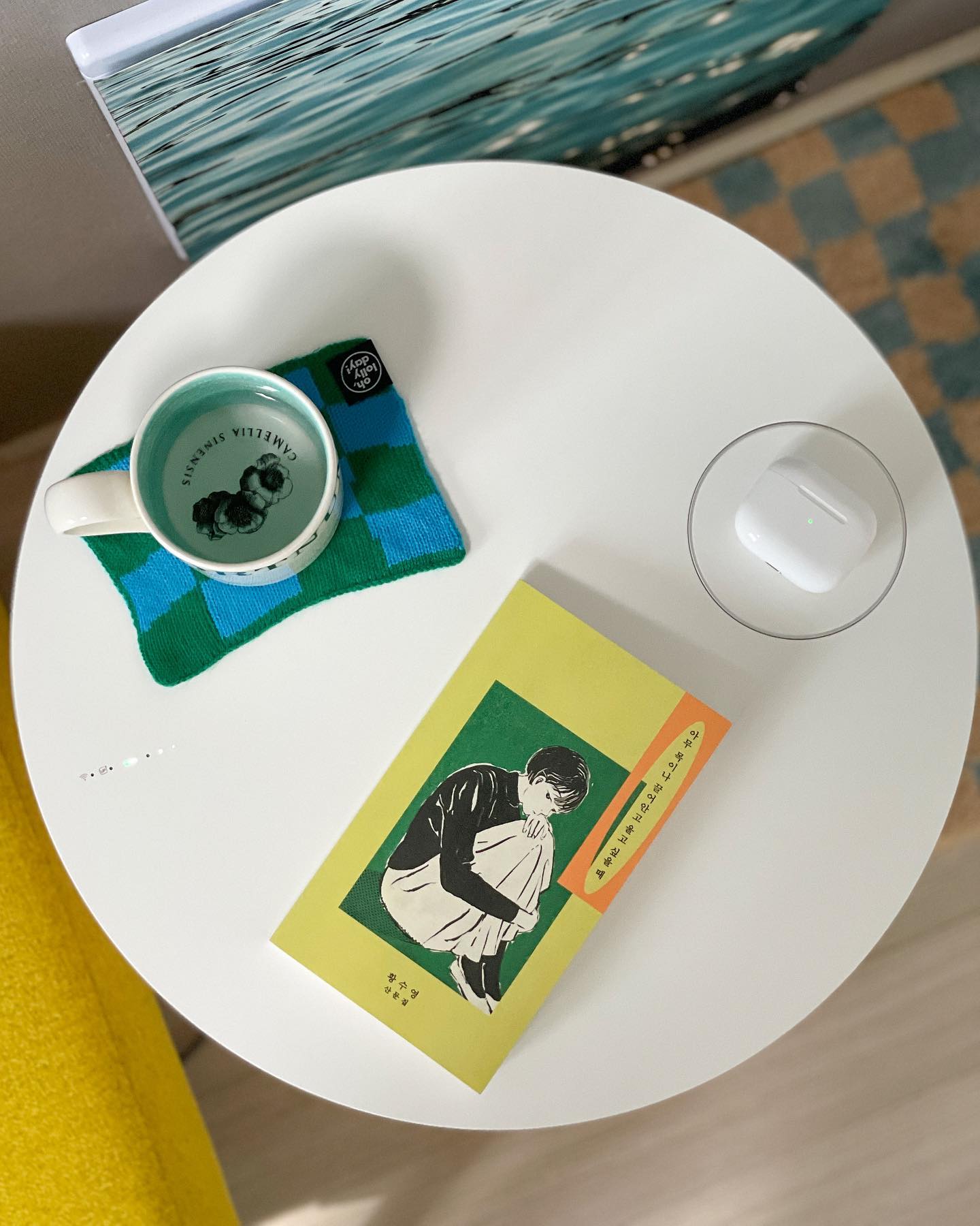 깔끔한 테이블 위에 책, 머그컵, 그리고 무선 이어폰이 놓여 있으며, 색감이 조화로운 코스터와 배경의 파란색 그림이 아늑한 분위기를 연출하는 공간