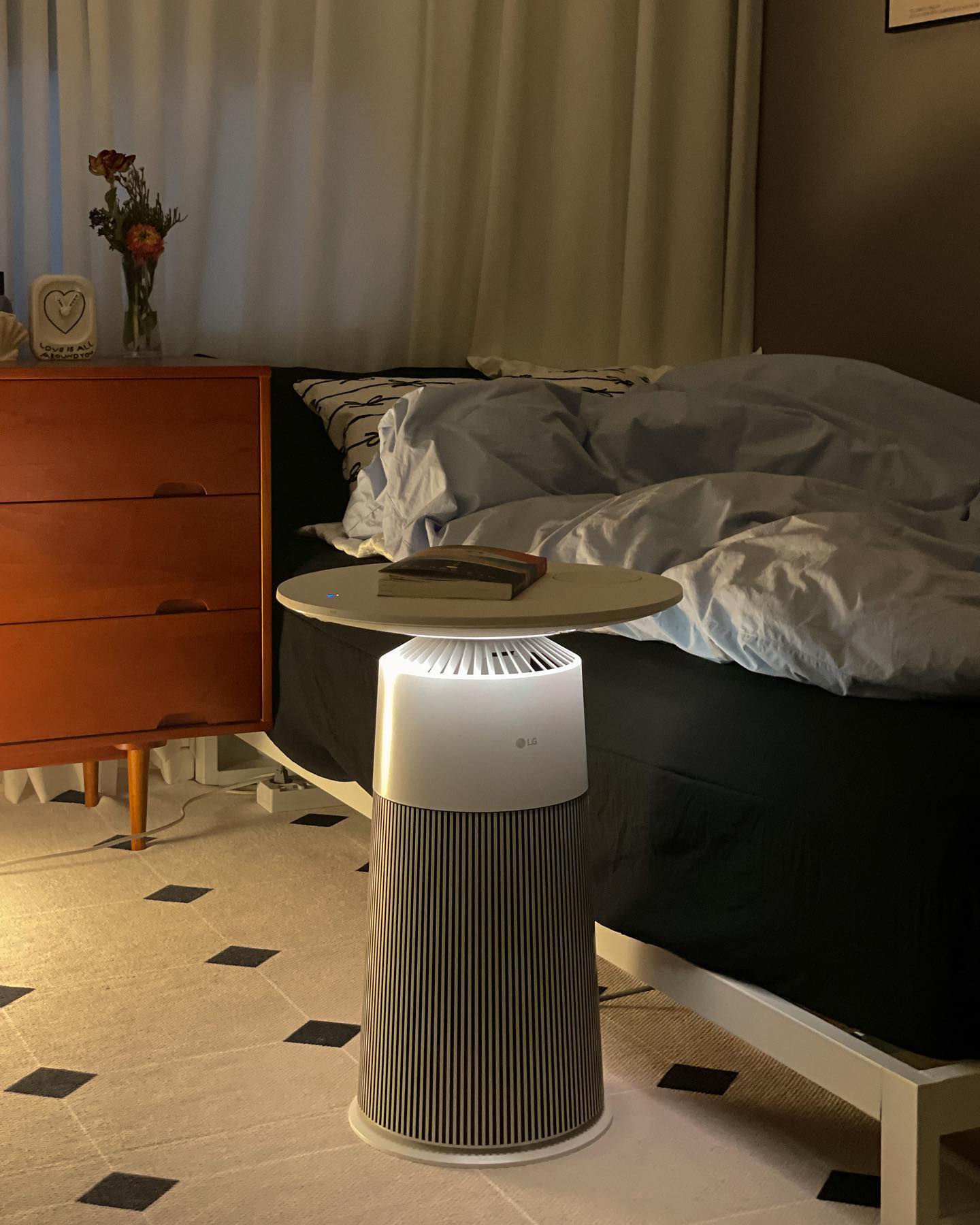 아늑한 침실 한쪽의 공기청정기 위에 책이 올려져 있는 모습. 침대 옆에 있는 공기청정기는 조명이 켜져 있으며, 침대와 나무 서랍장이 함께 보이는 따뜻한 분위기의 실내 장면