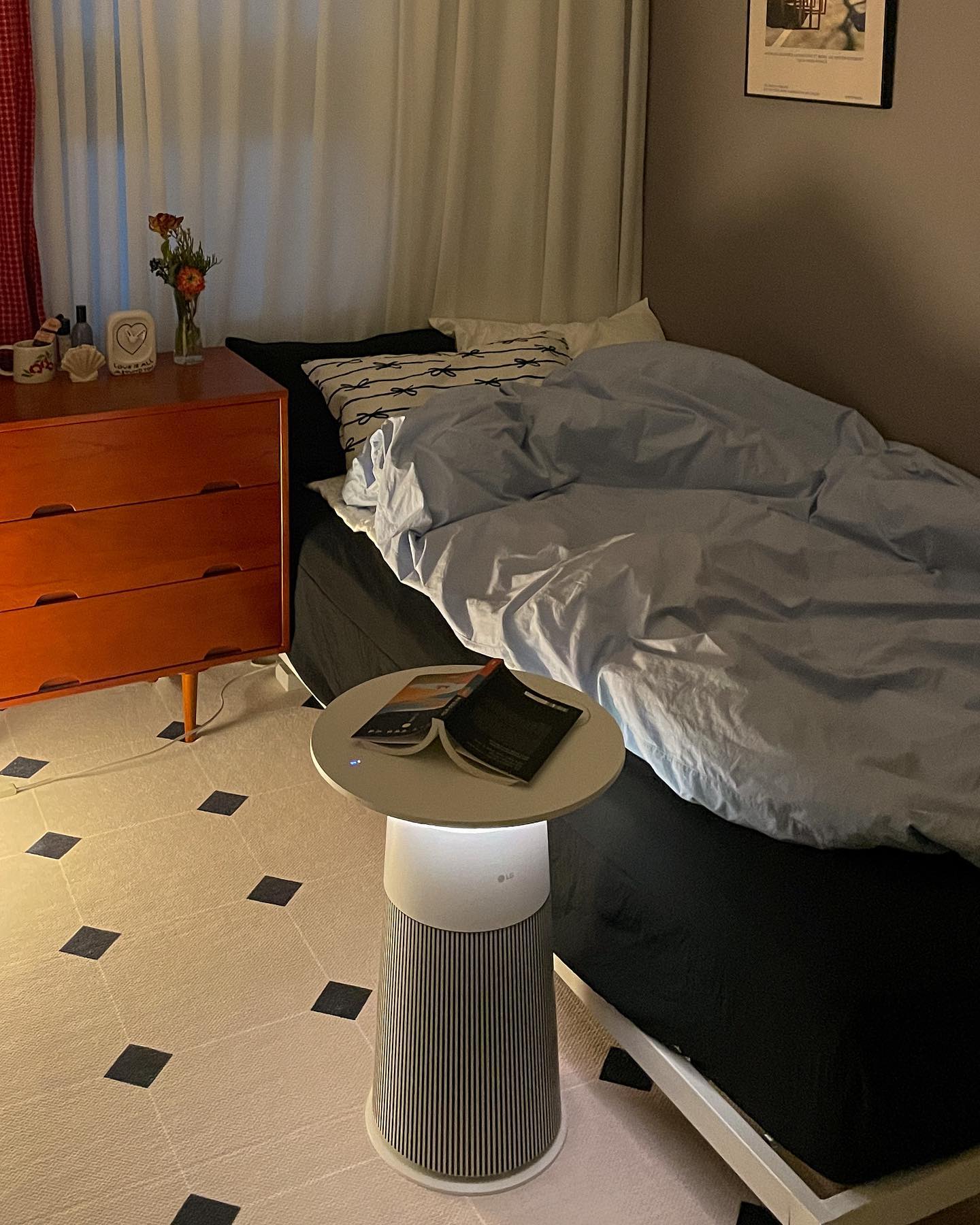 침실의 공기청정기 위에 펼쳐진 책이 놓여 있고, 침대와 나무 서랍장이 함께 보이는 아늑한 실내 장면