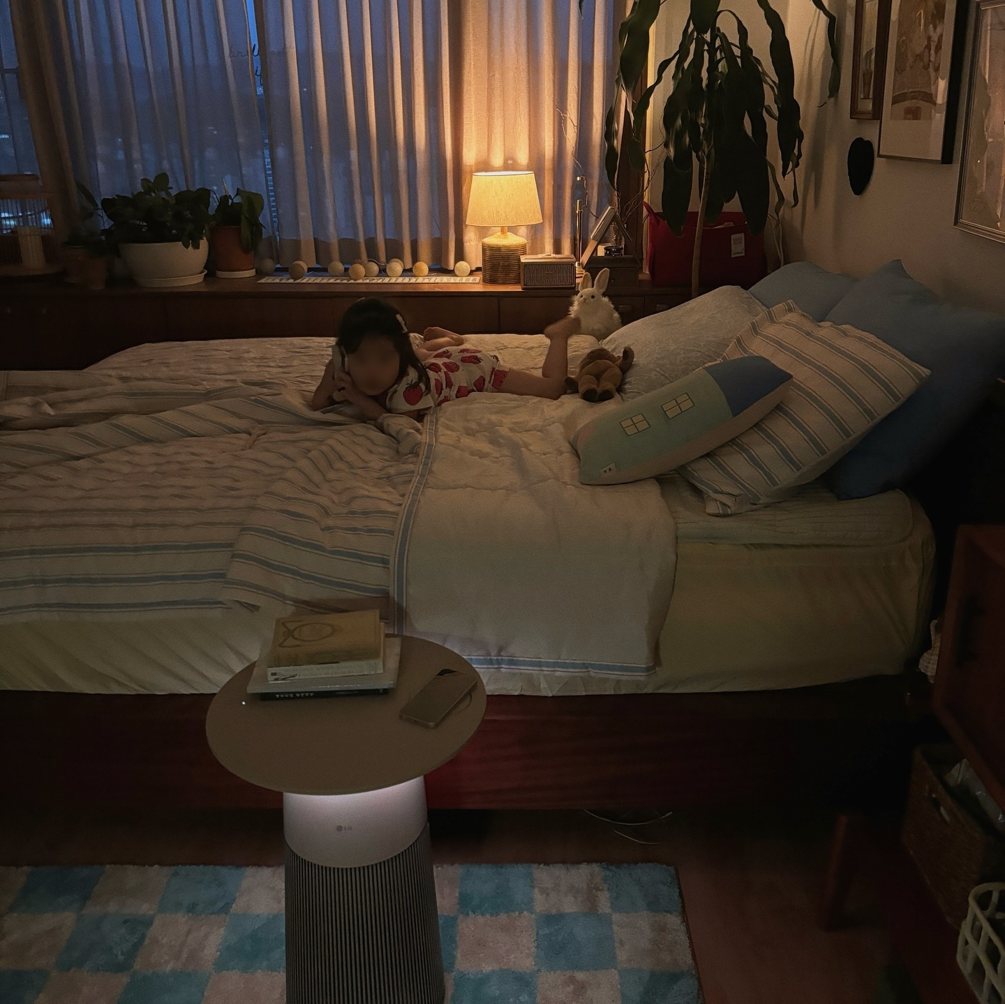 어두운 방 안, 테이블형 공기청정기와 조명들이 켜져 있음. 나무 프레임 침대 위에는 침구들과 쿠션이 놓여 있고. 한 아이가 엎드려 있음. 테이블 위에는 책과 스마트폰이 있으며, 창가에는 커튼 걸려 있음.
