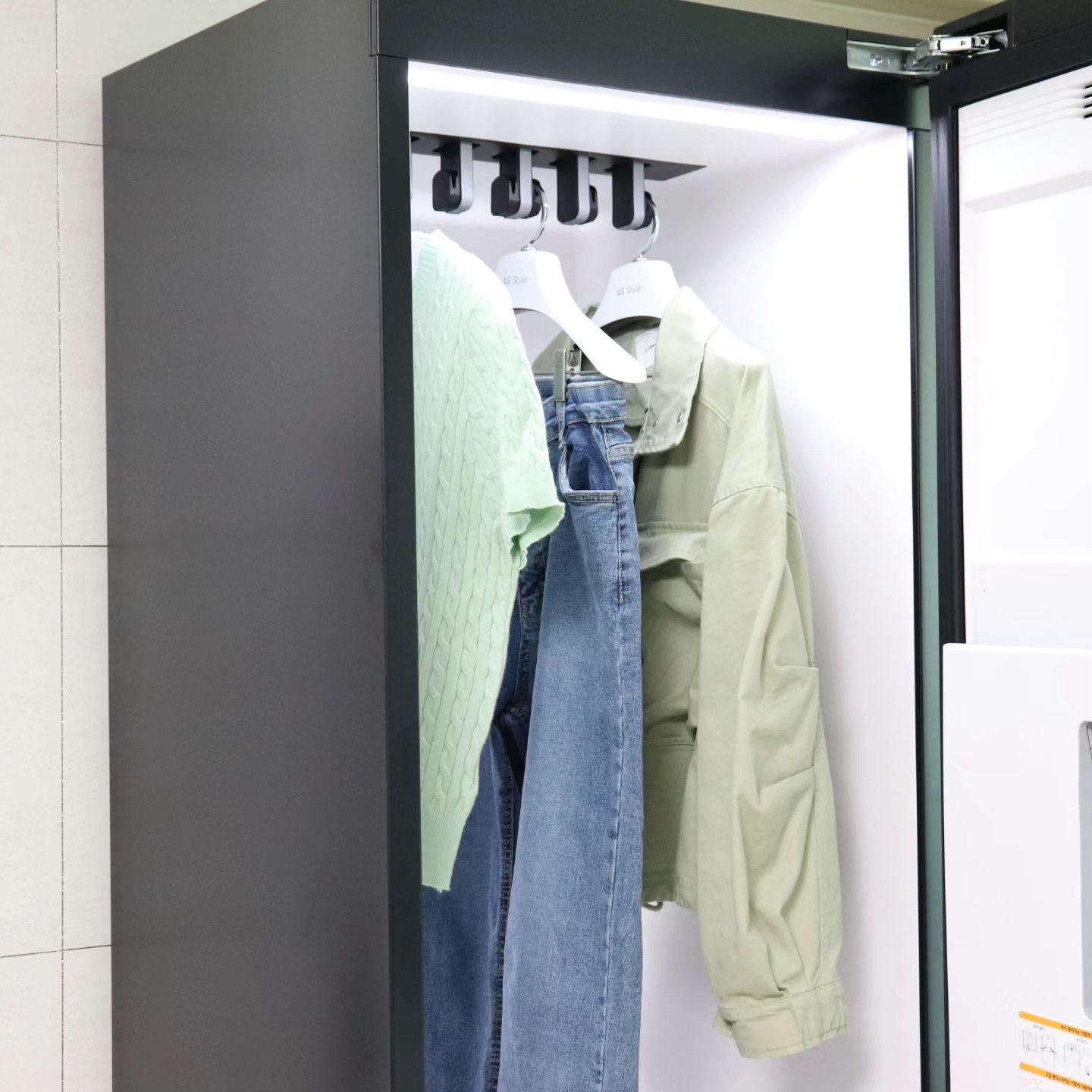 문이 열린 스타일러 내부에 연두색 니트, 청바지, 연두색 재킷이 옷걸이에 걸려 있고, 내부 조명이 밝게 켜져 있음.