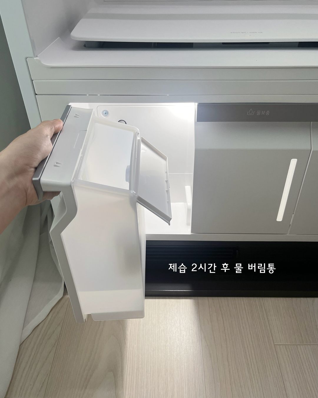 스타일러의 하단 물 버림통을 손으로 꺼낸 모습. 제습 2시간 후 물 버림통이라는 문구가 표시됨. 내부 조명이 켜져 있음.