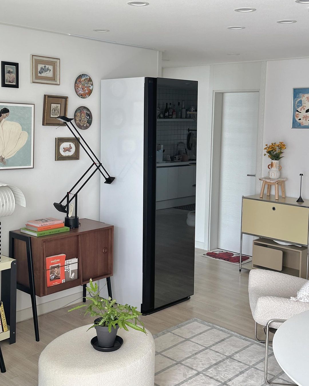 앞면은 블랙틴트 미러, 옆면은 화이트인 스타일러가 거실에 배치됨. 원형 테이블에 화분이 놓여있고, 벽에는 액자들이 걸려 있고, 우드 가구와 장식 소품들이 배치되어 있음.