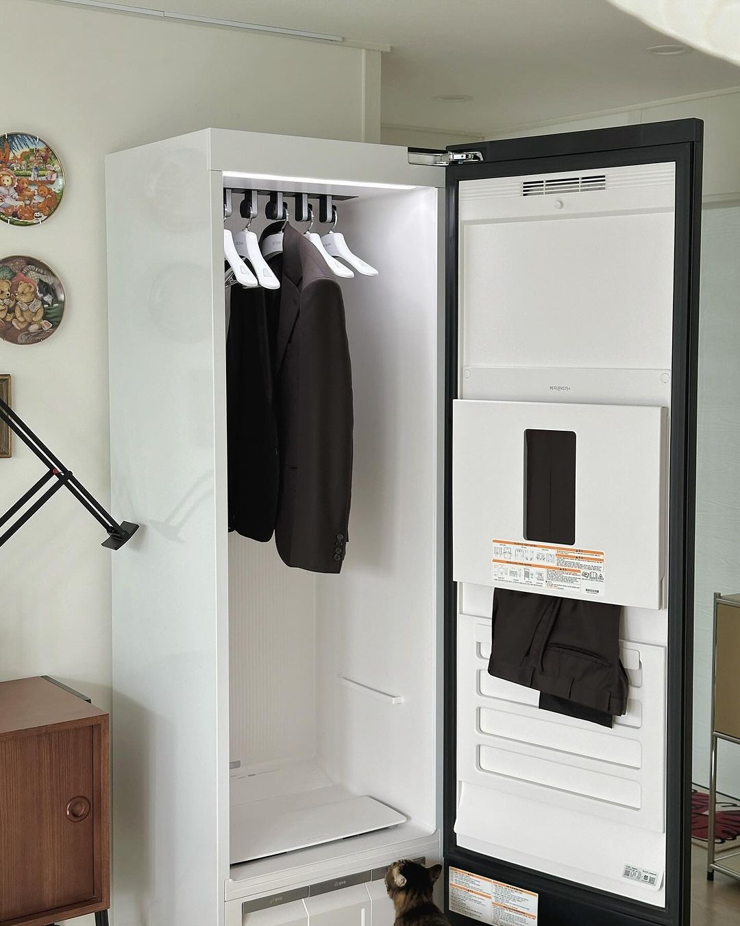 문이 열린 스타일러 내부에 검은색 정장 재킷과 바지가 걸려 있고, 조명이 켜짐. 하단에는 물통이 보이며, 고양이가 기기를 바라보고 있음. 배경에는 원목 수납장과 벽에 액자가 걸려 있음.