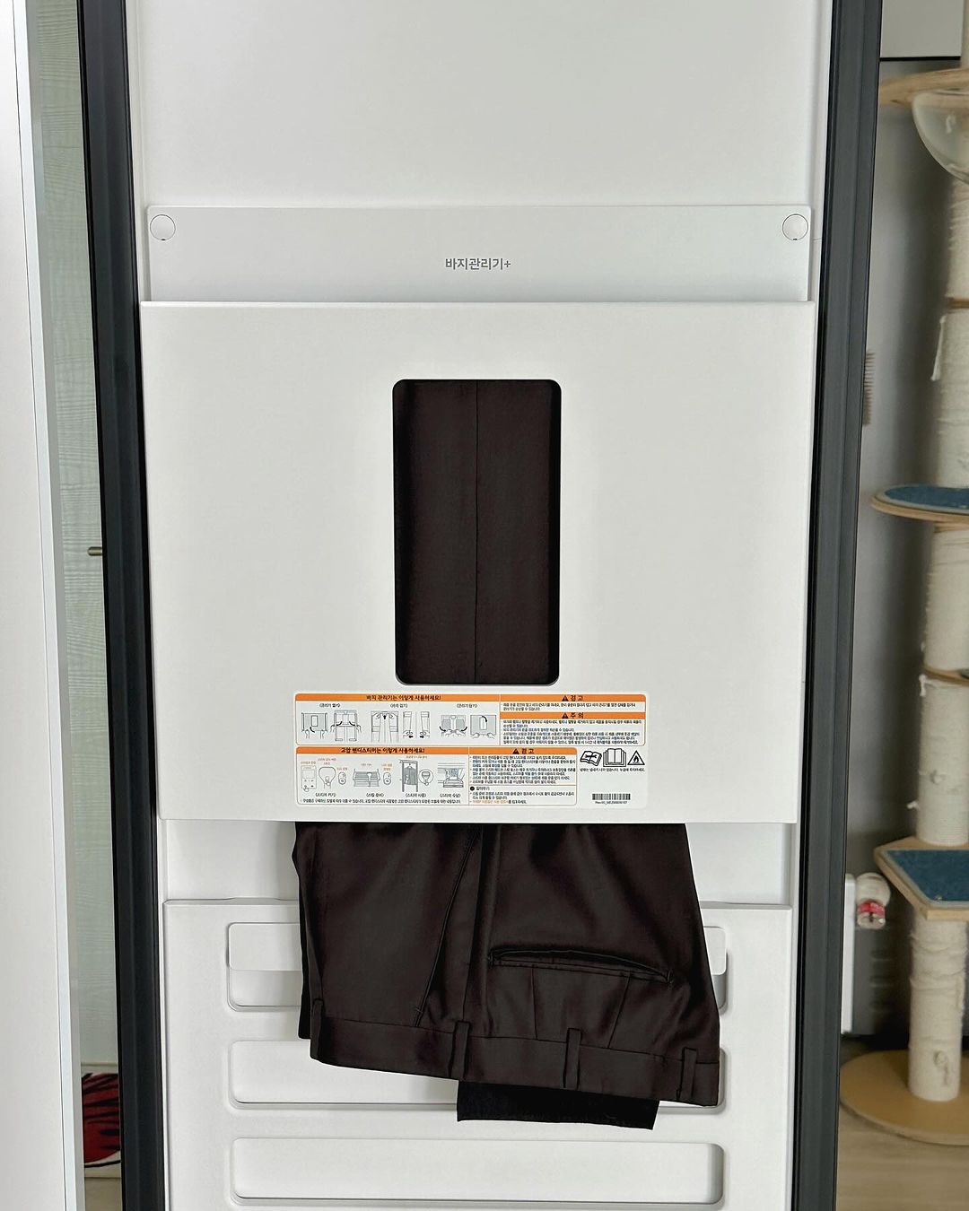 스타일러 내부 바지 관리기에 검은색 정장 바지가 걸려 있음. 관리기 문 안쪽에는 사용 안내 스티커가 부착되어 있음. 배경에는 캣타워가 보임.