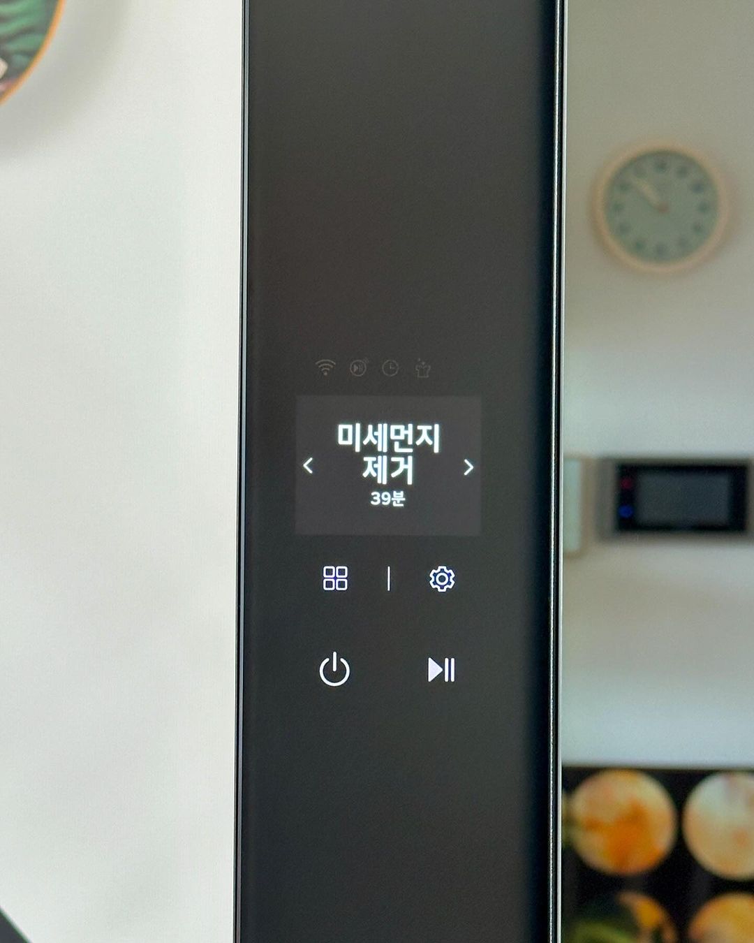 스타일러 전면 화면에 미세먼지 제거 39분 표시됨. 아래엔 네 개의 아이콘(메뉴, 설정, 전원, 일시정지) 버튼이 있고, 배경에는 벽시계와 TV가 있음.