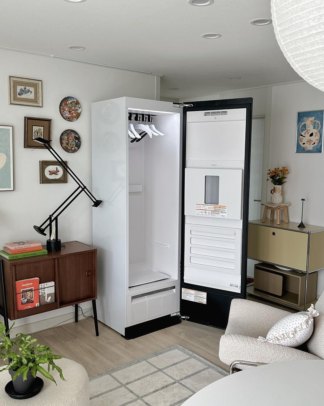 문이 열린 스타일러 내부에 옷걸이가 여러 개 걸려 있고, 조명이 밝게 비추고 있음. 문 안쪽에는 바지 관리기가 부착되어 있으며, 벽에는 액자들이 걸려 있고, 우드 가구와 장식 소품들이 배치되어 있음.