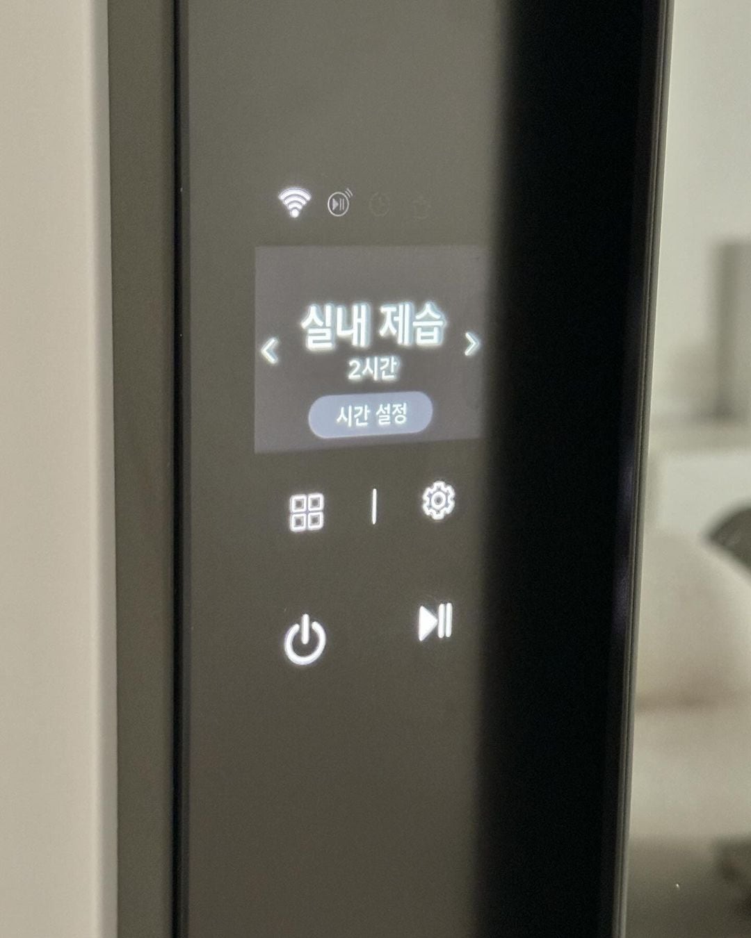 스타일러 전면 화면에 실내 제습 2시간 표시됨. 아래엔 시간 설정과 네 개의 아이콘(메뉴, 설정, 전원, 일시정지) 버튼이 있고, 상단엔 와이파이 외 기타 아이콘이 있음.