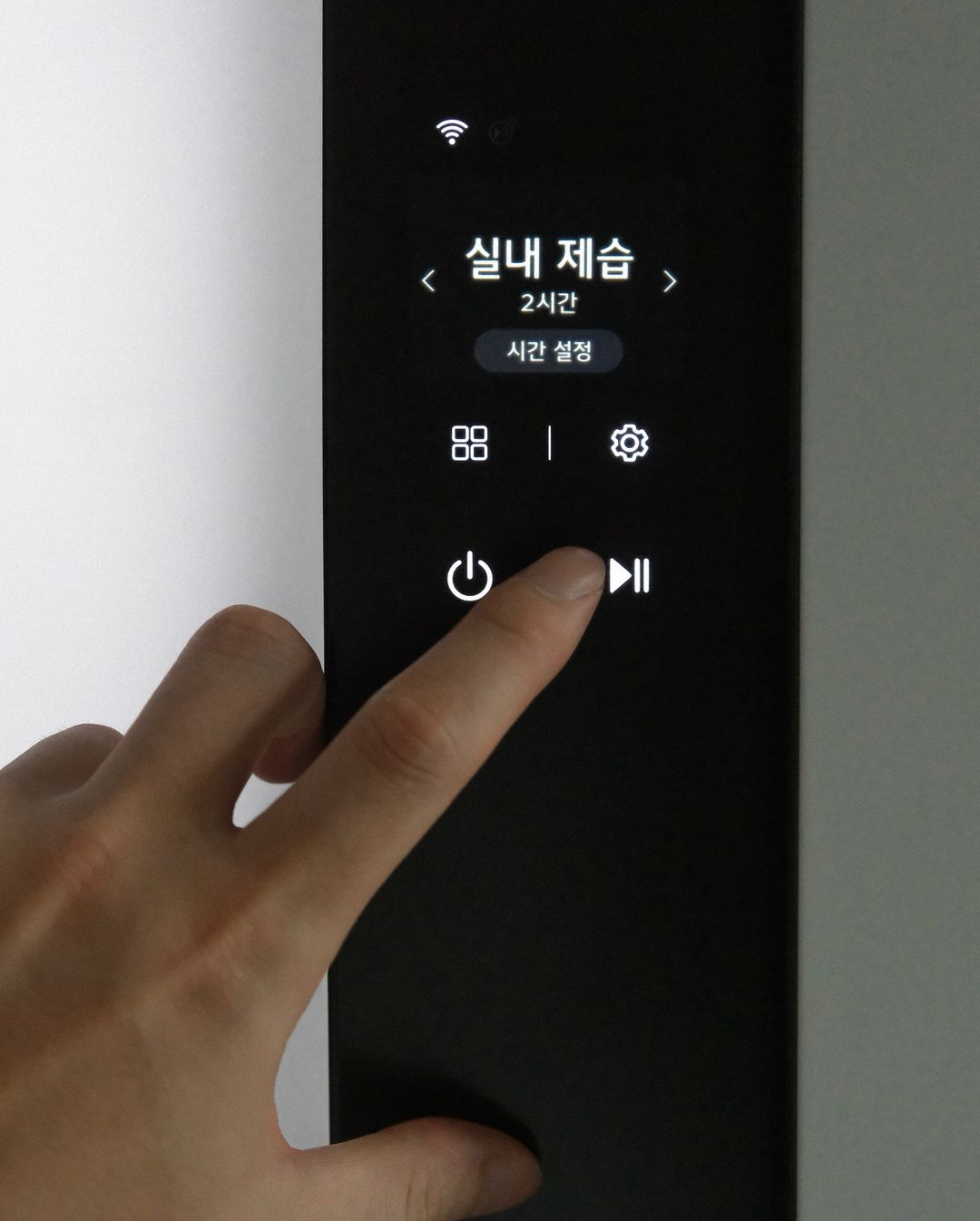 스타일러 전면 화면에 실내 제습 2시간 표시됨. 아래엔 시간 설정과 네 개의 아이콘(메뉴, 설정, 전원, 일시정지) 버튼이 있으며, 상단에는 와이파이 아이콘이 있음.