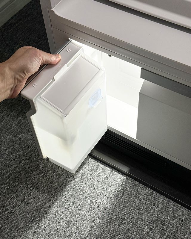 스타일러 하단에서 손이 물통을 꺼내고 있는 모습임. 투명한 물통에 뚜껑이 닫혀 있으며, 내부 공간이 밝게 빛나고 있음.