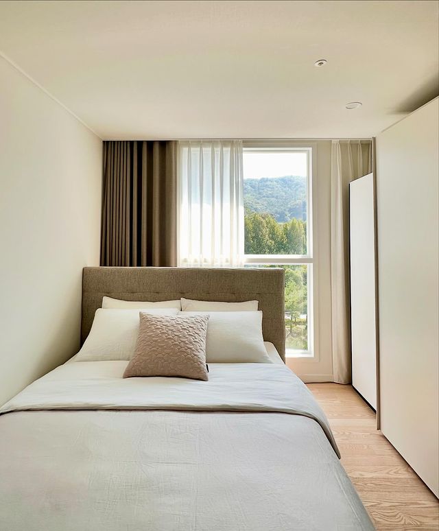 침대 위에 베이지색 쿠션과 흰색 침구가 놓여 있으며 창가에는 커튼이 걸려 있음. 오른쪽에는 화이트 스타일러과 붙박이장이 배치돼 있으며, 바닥은 우드 톤임. 창밖으로 푸른 산이 보임.