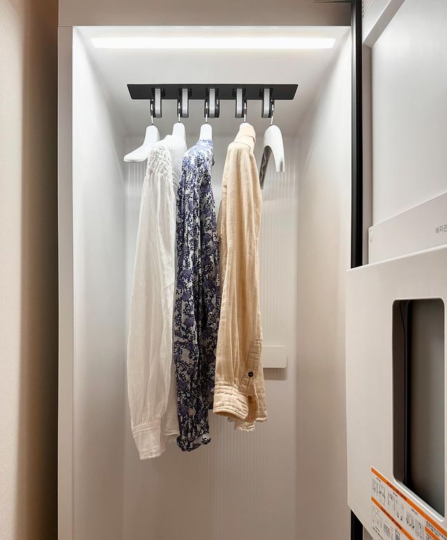 문이 열린 스타일러 내부에 흰색, 파란 패턴, 베이지색 셔츠가 옷걸이에 걸려 있고, 문 안쪽에는 바지 관리기 패널이 장착되어 있고, 내부 조명이 켜져 있음.