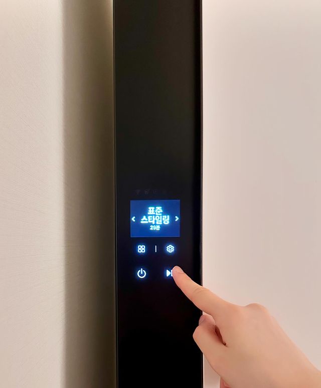 스타일러 디스플레이 화면에 표준 스타일링 29분 이 표시됨. 네 개의 아이콘(메뉴, 설정, 전원, 일시정지) 버튼이 있으며, 한 손이 시작 버튼을 누르고 있음.