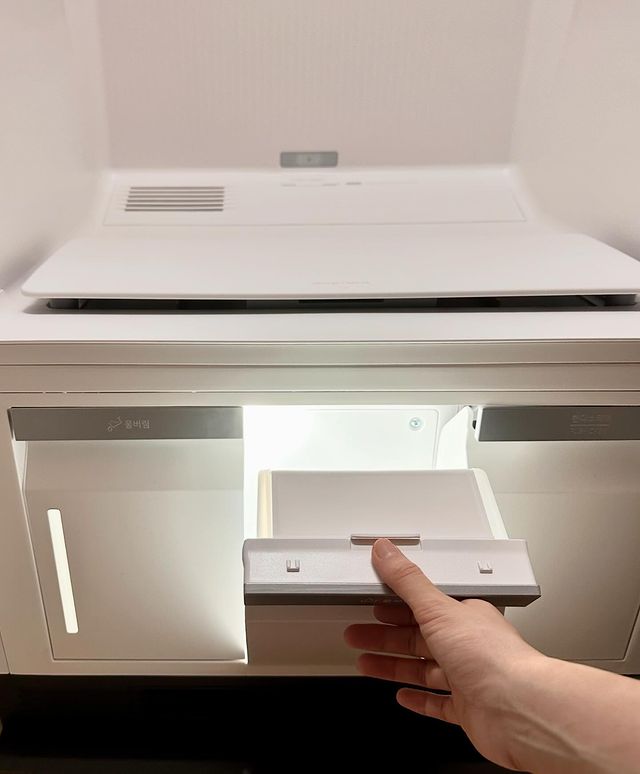 스타일러 하단에서 손으로 작은 물통을 꺼내는 모습. 내부 조명이 켜져 있음.