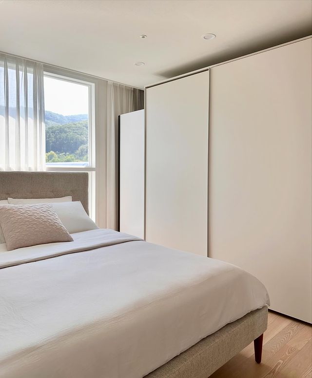 화이트 톤의 침실에 창가 커튼이 반쯤 드리워져 있으며, 침대 위엔 쿠션이 놓여 있음. 우측에는 화이트 스타일러과 붙박이장이 배치됨. 창밖으로 푸른 산이 보임.