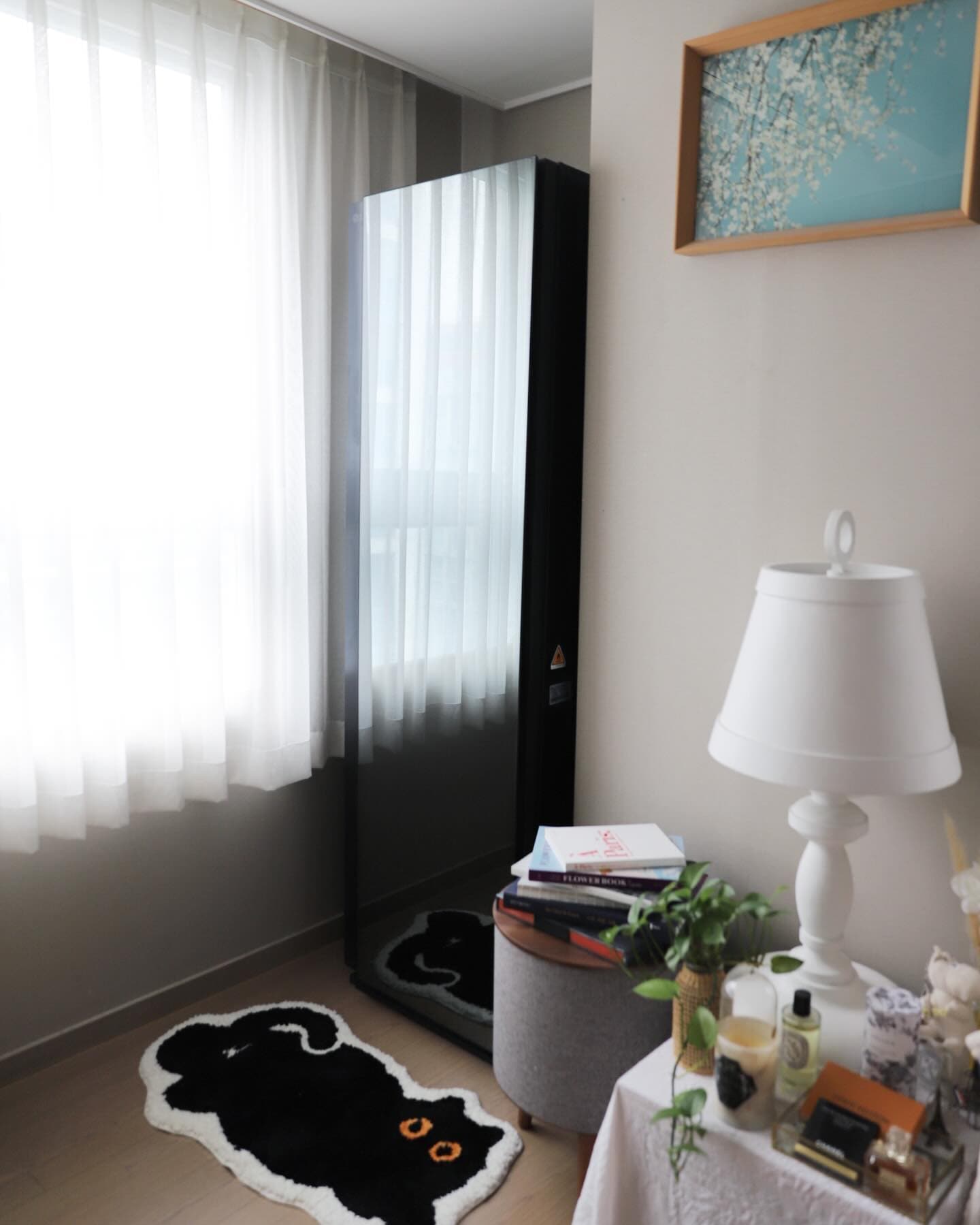 앞면이 블랙틴트 미러인 올블랙 색상의 스타일러가 창가에 놓여있음. 반사된 커튼과 바닥의 검은 고양이 러그가 보이며, 옆에는 원목 사이드 테이블 위에 책과 화분이 있음. 배경에는 스탠드 조명과 향수, 장식품 등이 있음.