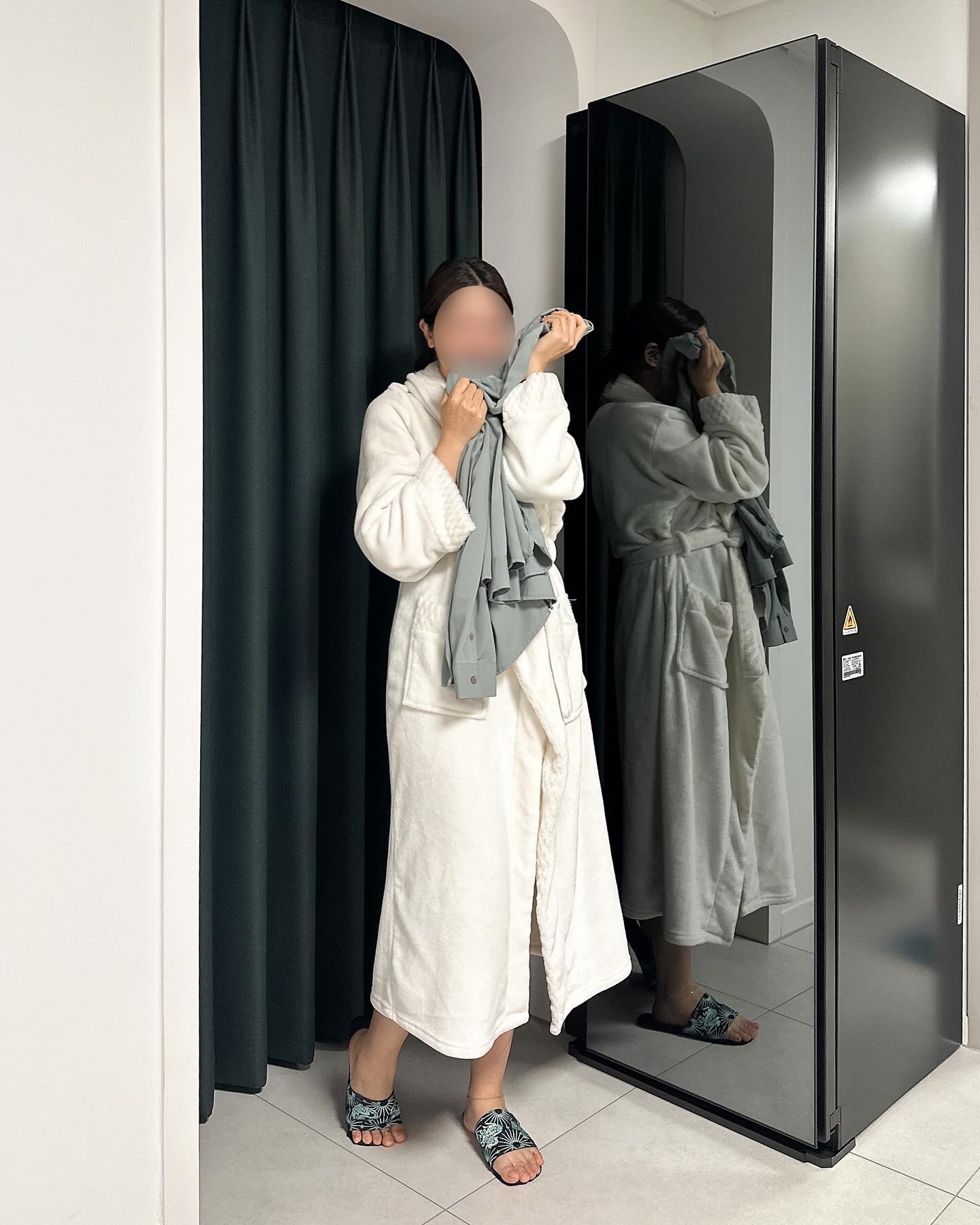 가운을 입은 여성이 앞면이 블랙틴트 미러인 올블랙 색상의 스타일러 앞에서 꺼낸 옷을 얼굴에 대고 냄새를 맡고 있음. 배경에는 어두운색 커튼이 있음.