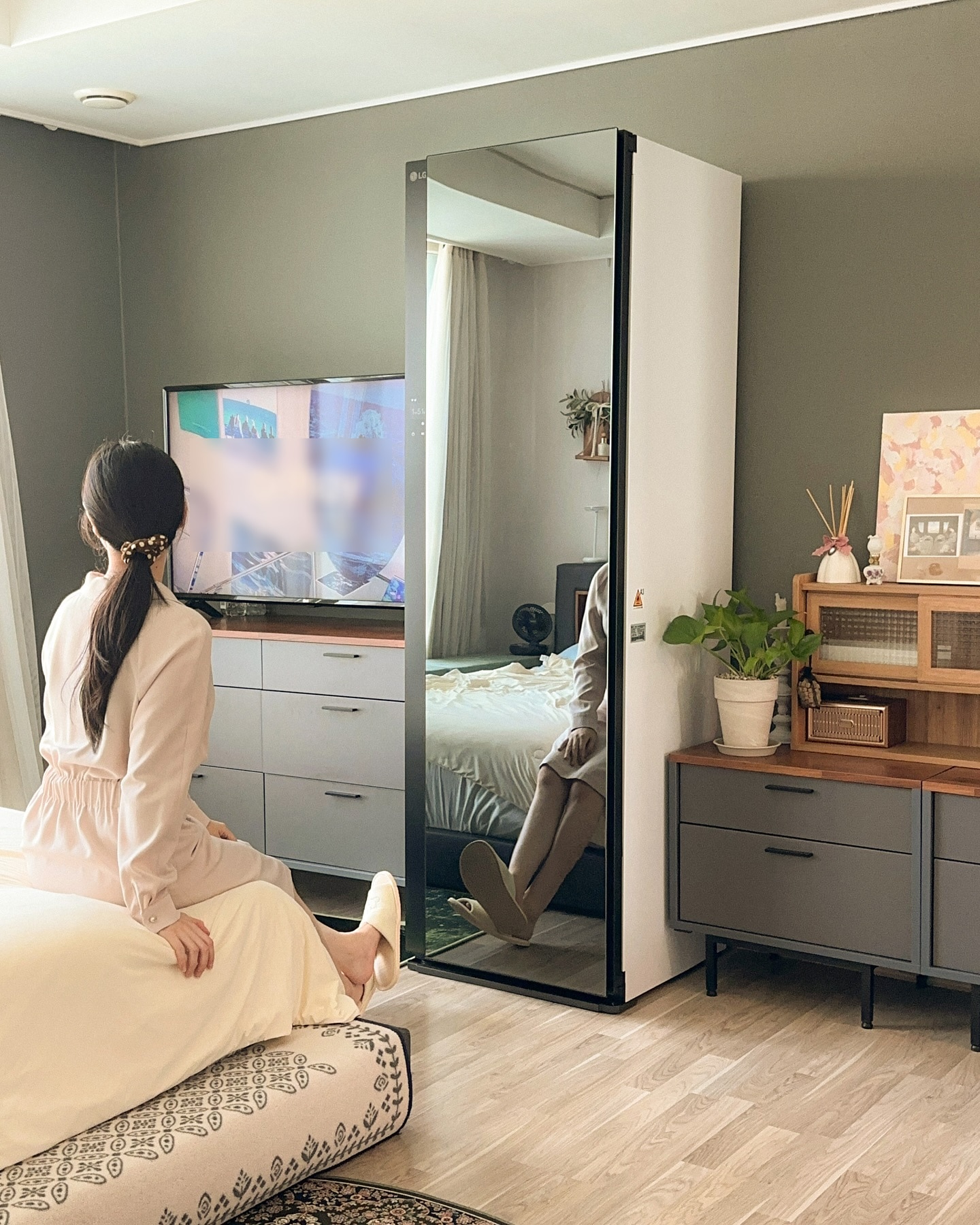 침대에 앉아 TV를 보는 여성, 맞은편에 앞면은 블랙틴트 미러, 옆면은 화이트인 스타일러가 있음. 스타일러 거울에는 여성의 모습이 반사됨. 옆에는 서랍장과 장식장이 있으며, 화분과 소품이 올려져 있음.
