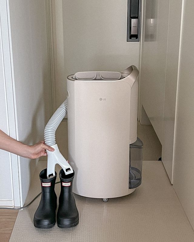 화이트 톤 중문이 보이는 현관 바닥에 휘센 제습기가 놓여 있음. 호스를 연결해 검은색 헌터 레인부츠 내부를 건조하는 모습임.