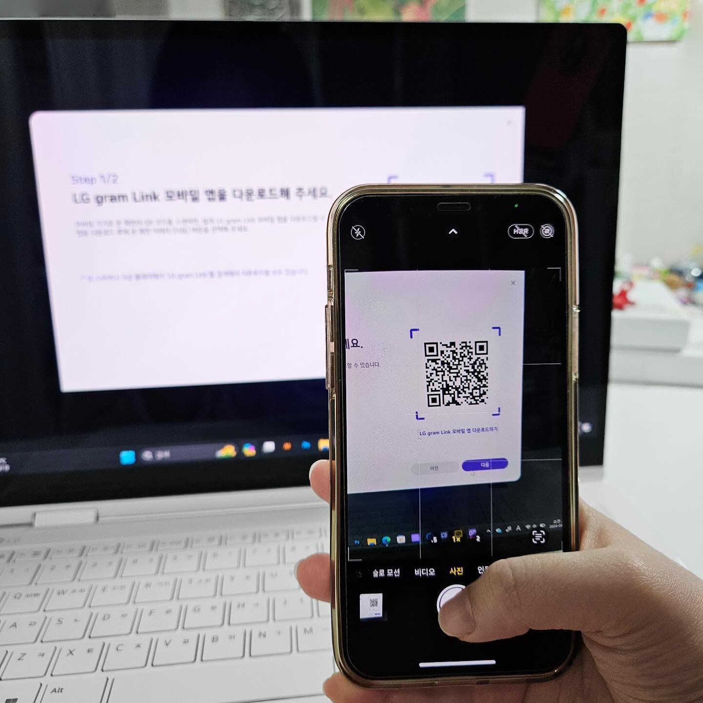스마트폰을 손에 든 사용자가 화면에 QR코드 앱을 실행, 뒤로는 LG 그램 노트북 에서 LG gram Link 모바일앱 다운로드 사이트 화면이 배경으로 보임.