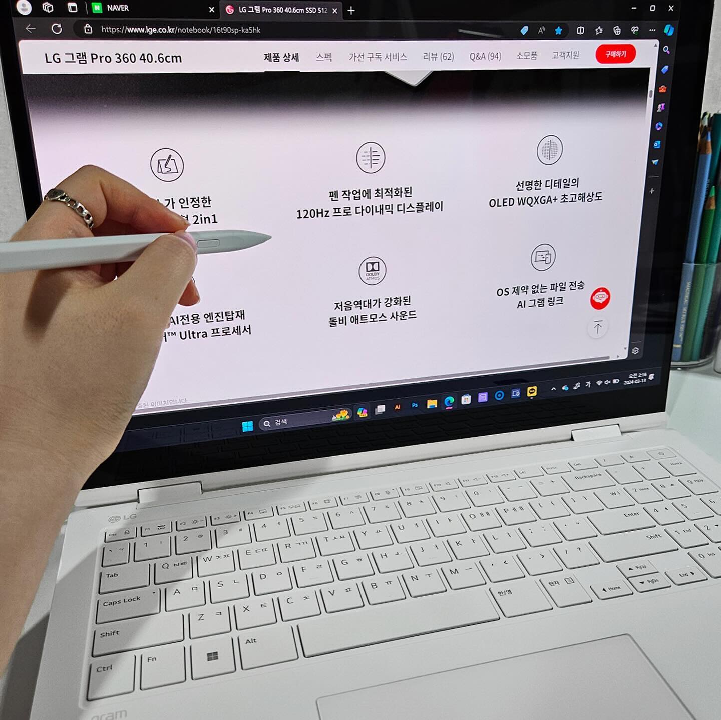 펜을 사용하여 터치스크린 기능을 활용하는 흰색 노트북 화면에 LG 그램 Pro 360의 제품 정보가 표시됨. 배경에는 필기 도구가 담긴 투명 용기가 놓여 있음.