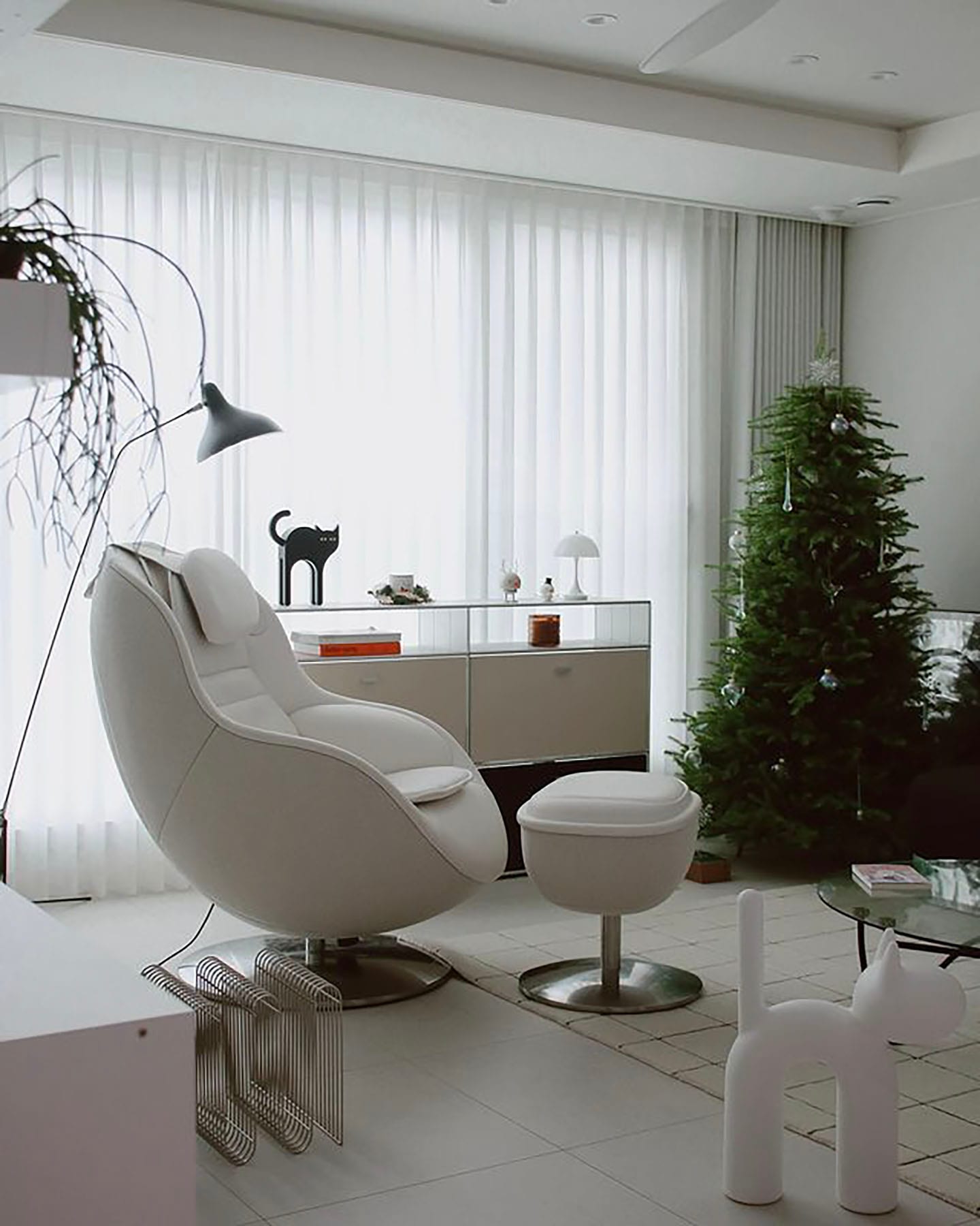 따뜻한 분위기의 거실 공간으로, 커튼이 드리워진 창가 옆에 편안한 디자인의 화이트 안마의자가 놓여 있으며, 한쪽에는 크리스마스트리가 장식된 모습